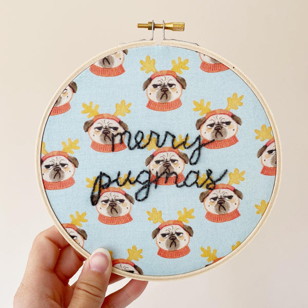 hand-stitched embroidery with text that reads "merry pugmas" on fabric with grumpy pugs wearing reindeer ears